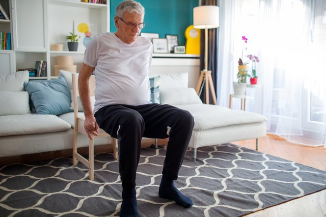 An elderly man performing balance exercises with the aid of a chair.